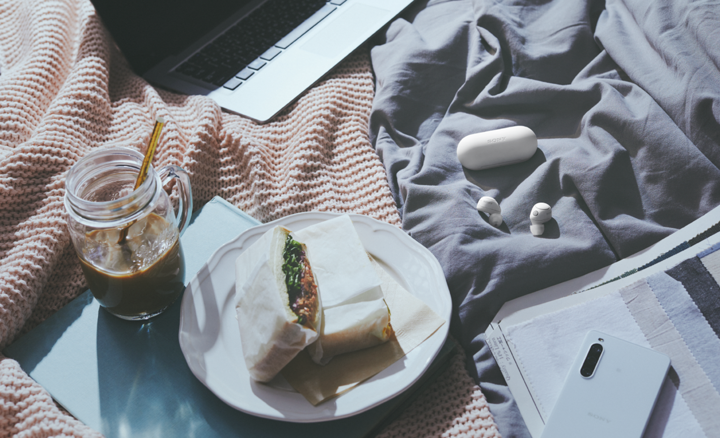 Abbildung der weißen WF-C700N Kopfhörer mit Etui auf einer Picknickdecke mit Essen, Getränk, einem Laptop und einem Handy